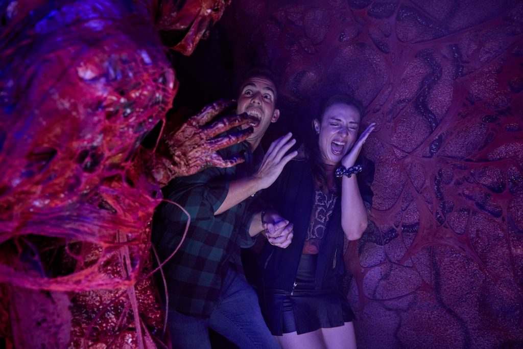 A couple holding hands and screaming in terror as a slimy creature reaches its decaying hand out toward them in the darkness.